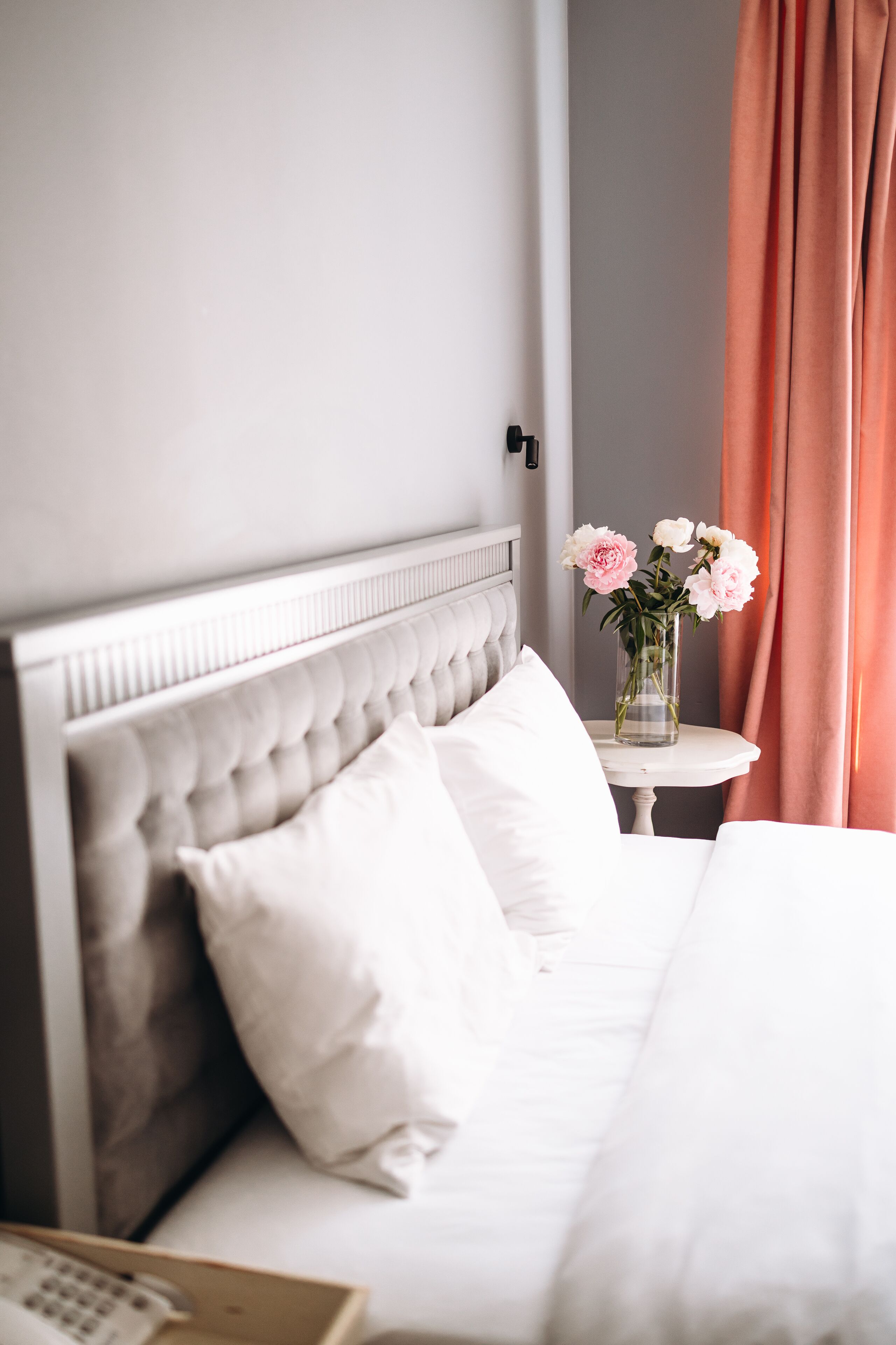 Una cama bien hecha con ropa blanca y un cabecero capitonado, un jarrón con flores sobre una mesita de noche y vista del interior de la habitación.