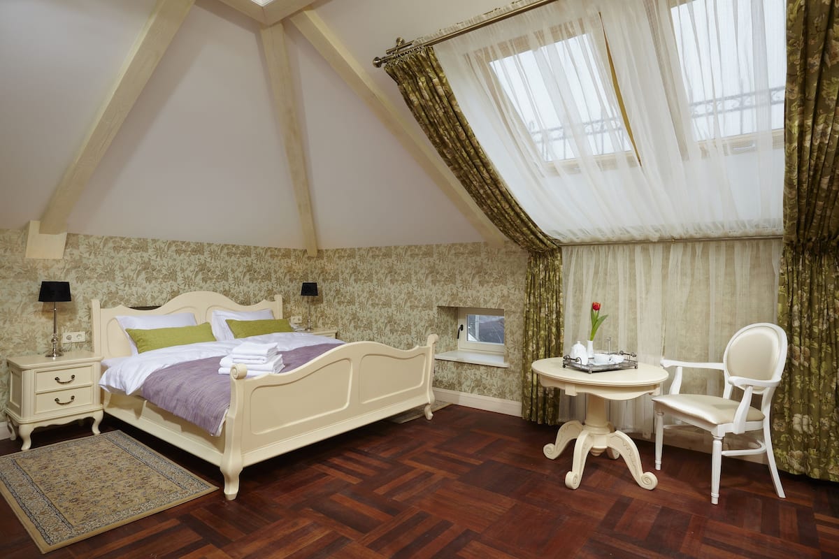 Une chambre à coucher avec un lit, des tables de chevet, une petite table avec une chaise, une fenêtre avec des rideaux et une vue sur l’extérieur.