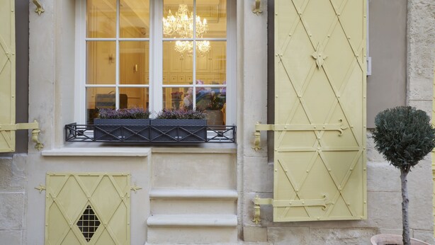 Eine Gebäudefassade mit Fenster, gelben Fensterläden, einem kleinen Balkon mit Pflanzen und einer Tür mit schmiedeeisernem Zierdekor.