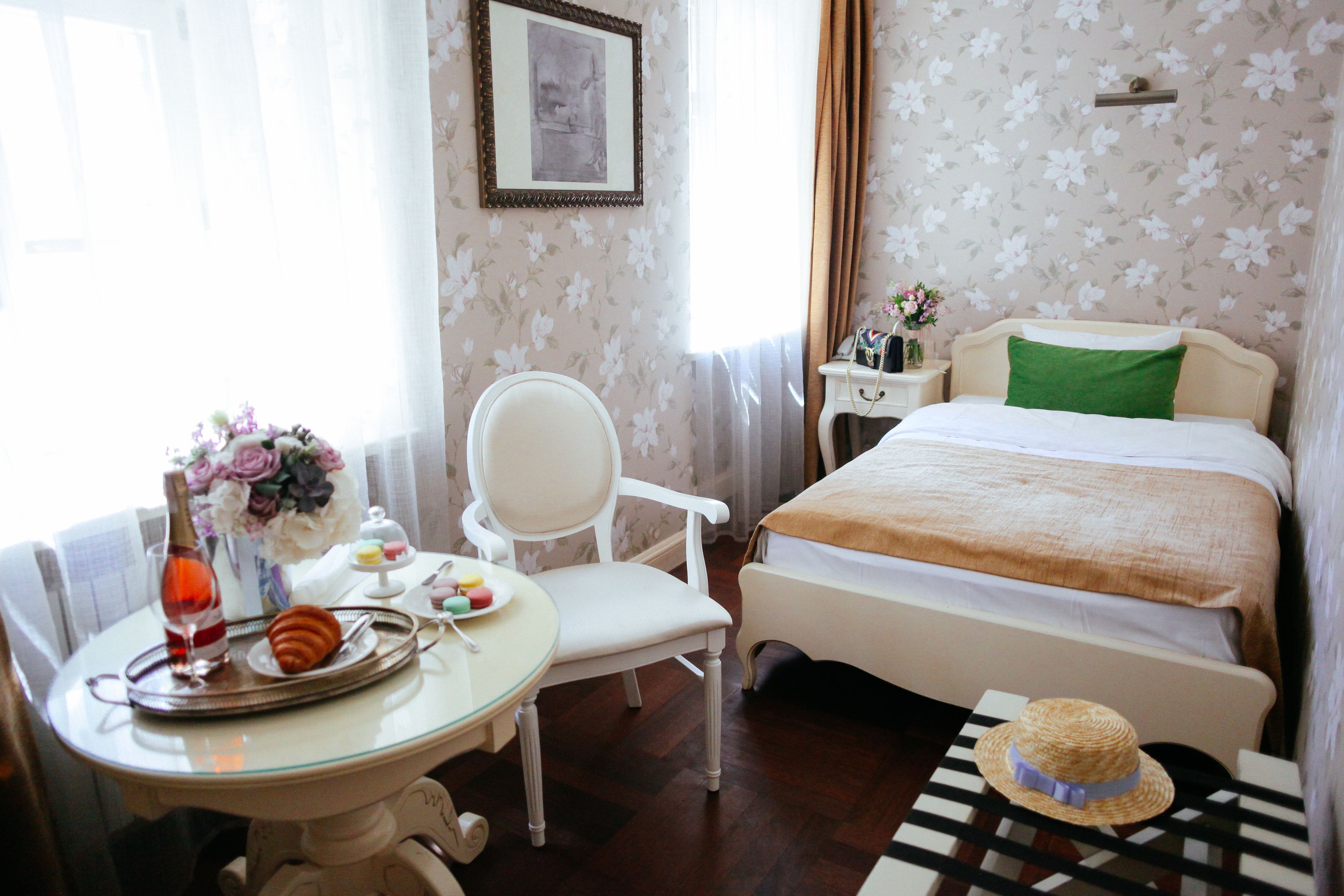 A hotel room with a bed, a chair, a small table with a tray of food, and a vase of flowers.