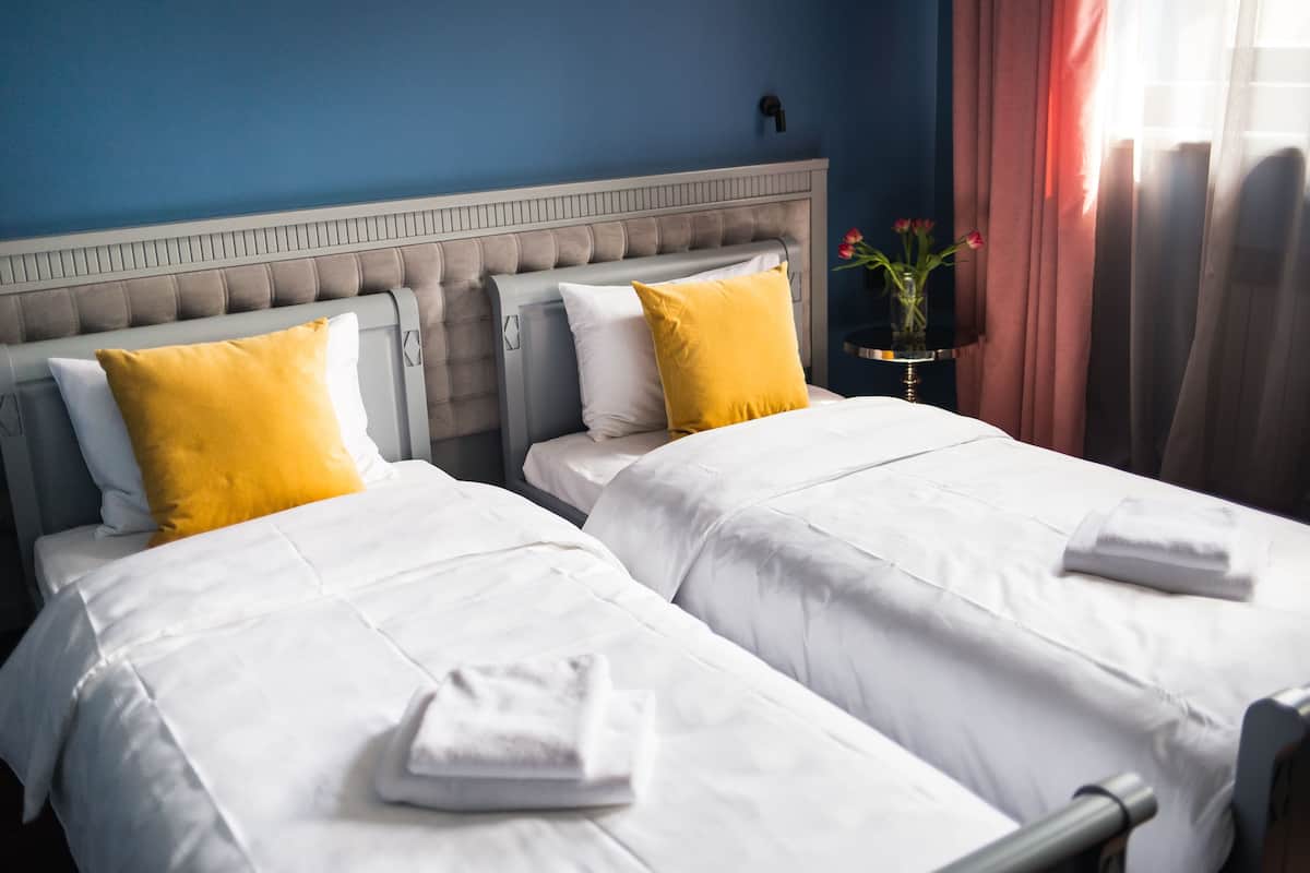 Une chambre d’hôtel avec deux lits, chacun étant équipé de draps blancs et d’un oreiller jaune. On y trouve une petite table sur laquelle est posé un vase de fleurs et une lampe.