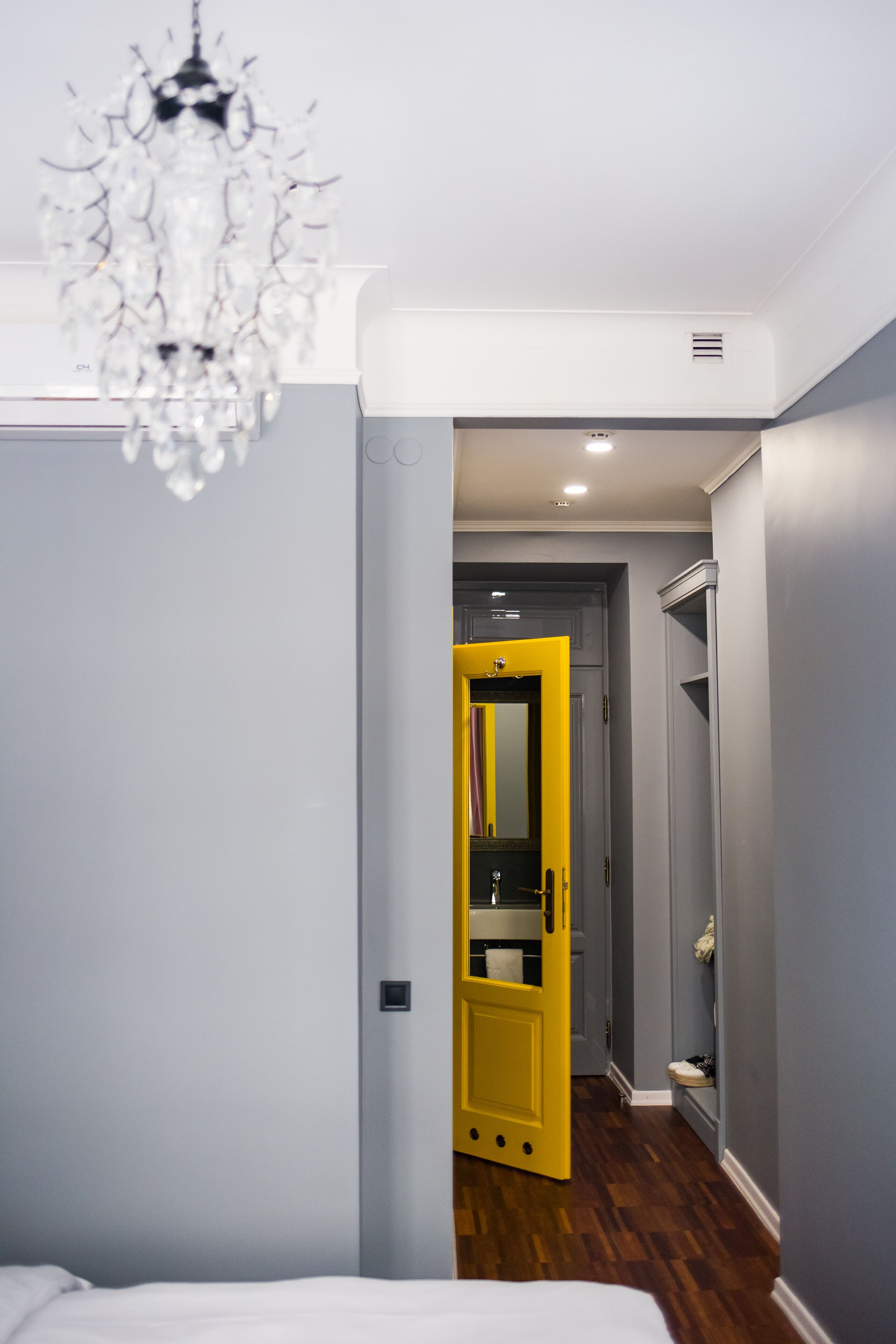 A hotel room with a chandelier, a bed, a bathroom with a yellow door, and a hallway with a mirror.