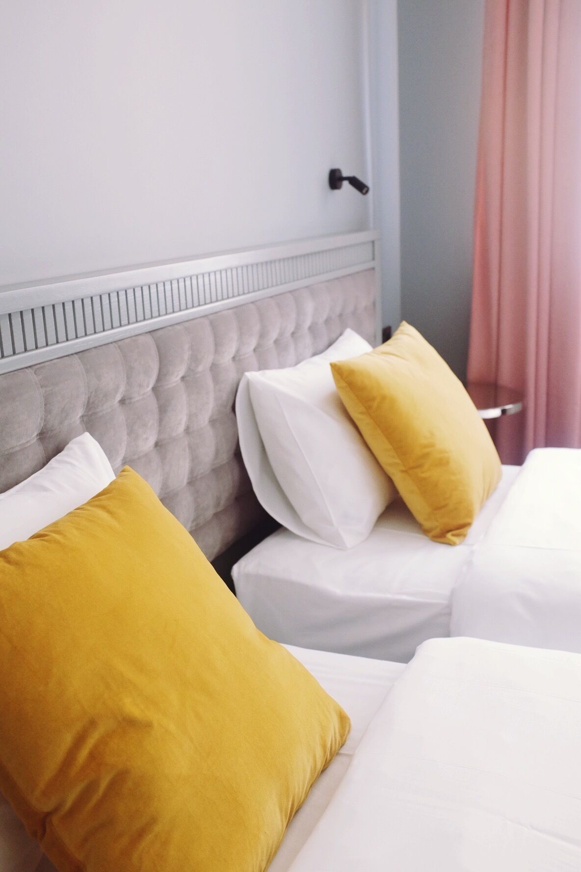 A hotel room with a tufted headboard, two beds with white linens, and a single yellow pillow on each bed.