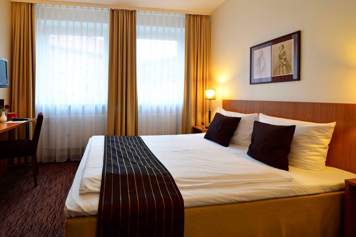 A hotel room with a large bed, a desk with a chair, a lamp, and a framed picture on the wall.