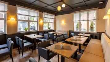 Un restaurant amb diverses taules i seients, grans finestres amb cortines i làmpades penjades a la paret.