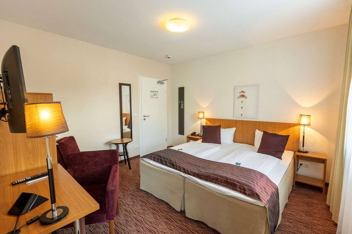 A hotel room with a bed, two bedside tables, a desk with a lamp, a chair, and a television.