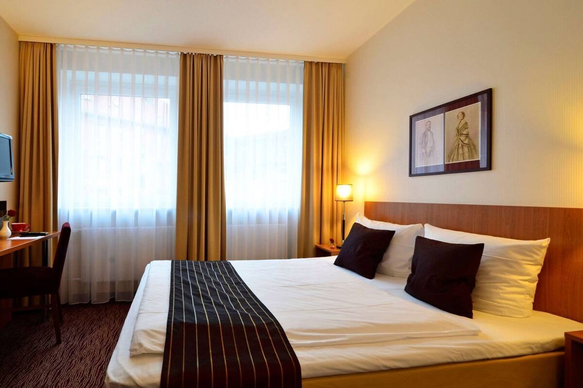 A hotel room with a bed, a desk, a chair, a lamp, and a framed picture on the wall.