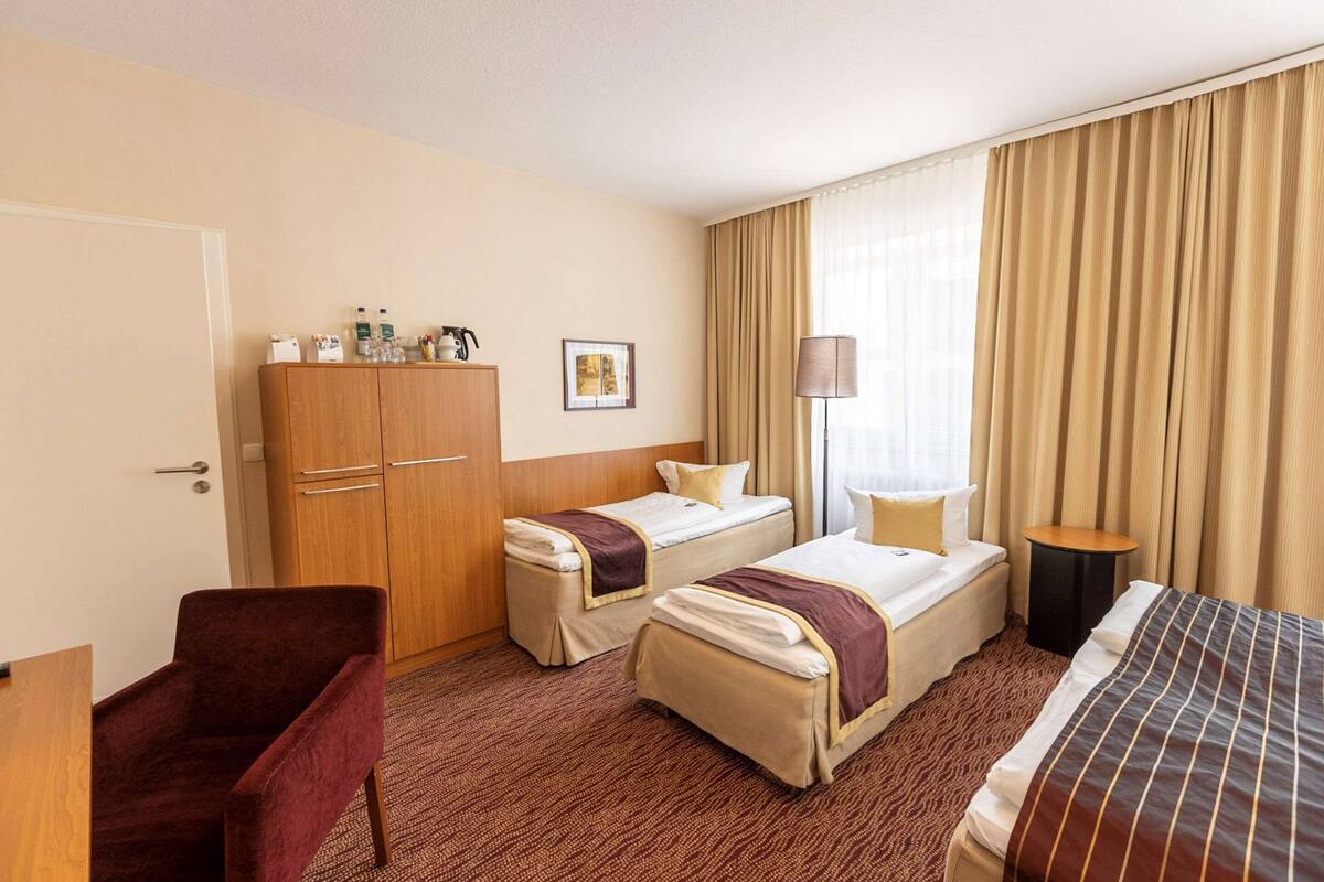 A hotel room with two single beds, a wooden wardrobe, a desk with a chair, and a large window with curtains.