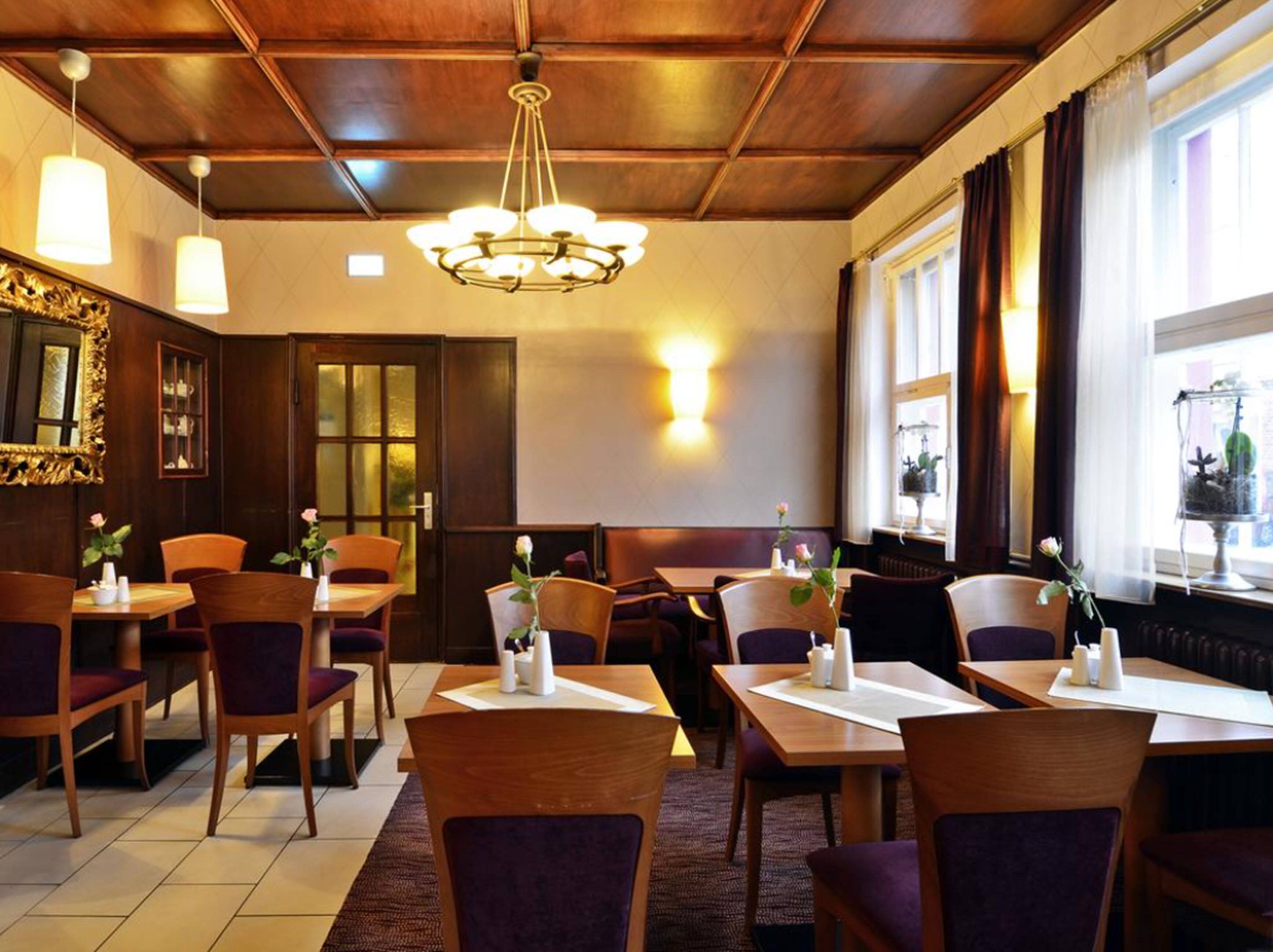 A restaurant with wooden tables and chairs, a chandelier, and a window with curtains.