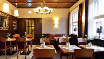 A restaurant with wooden tables and chairs, a chandelier, and a window with curtains.