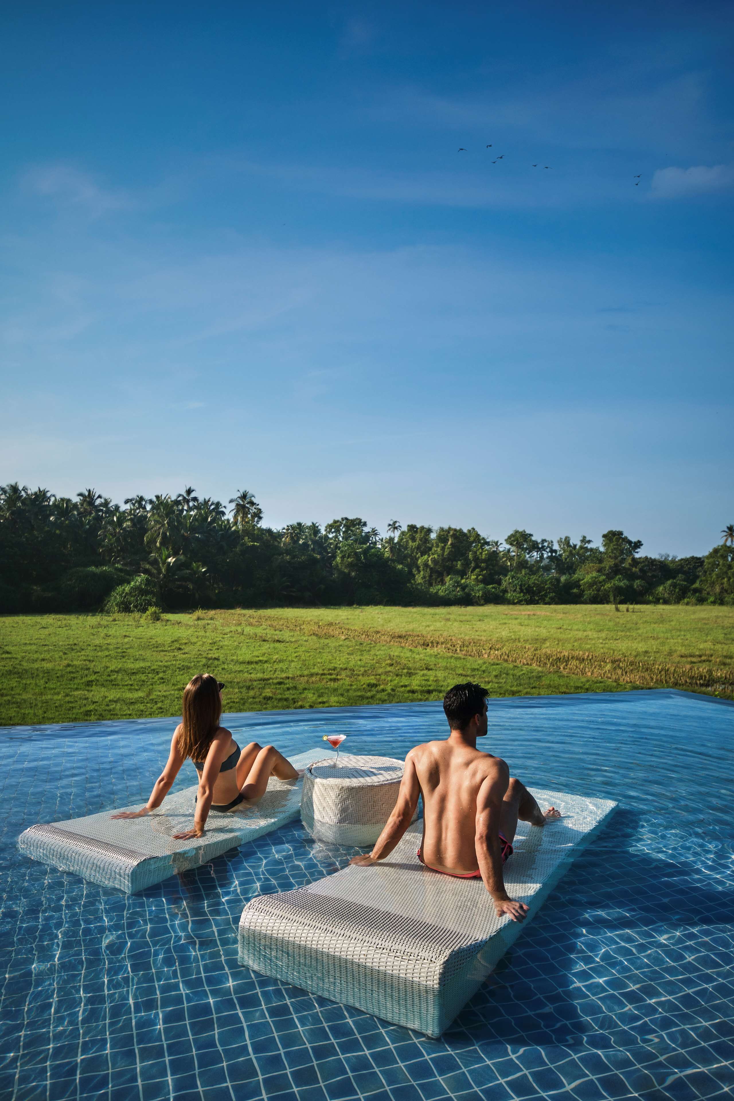 Deux personnes se détendent sur des chaises longues flottantes dans une piscine, d’où elles ont une vue dégagée sur un champ verdoyant et des arbres en arrière-plan.