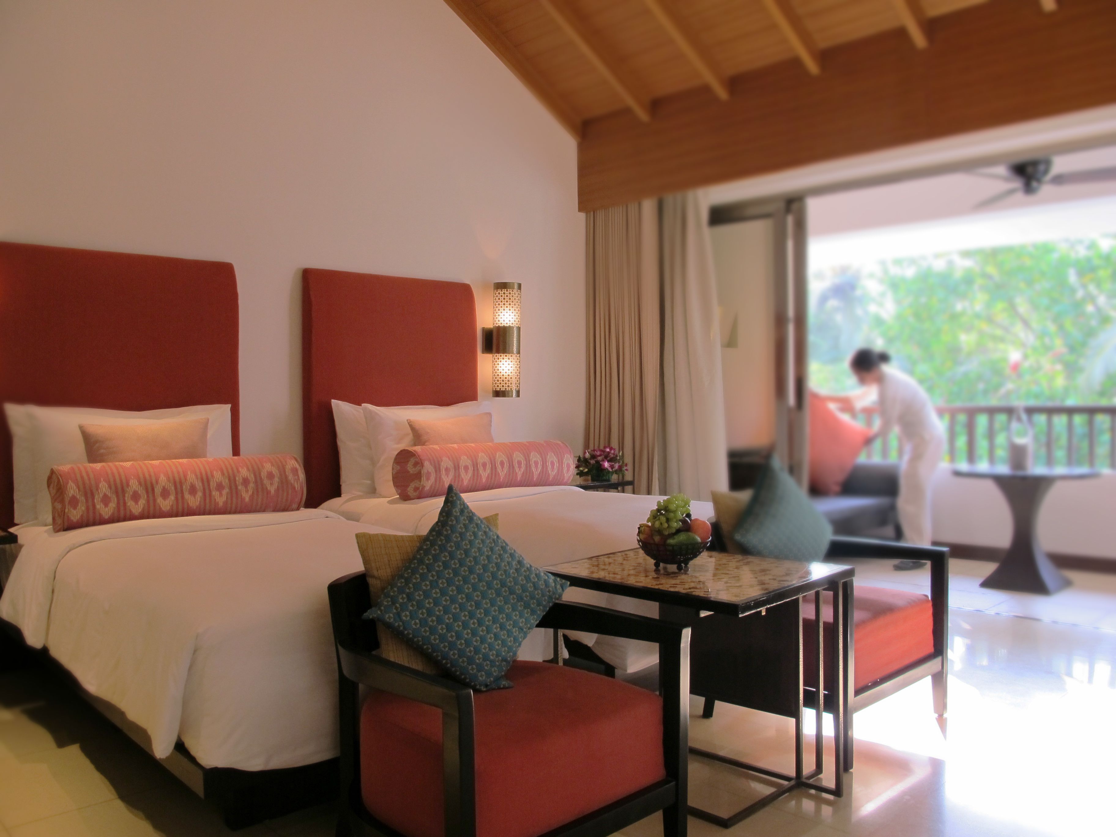A hotel room with two beds, a small table with a fruit bowl, and a balcony with a person outside.