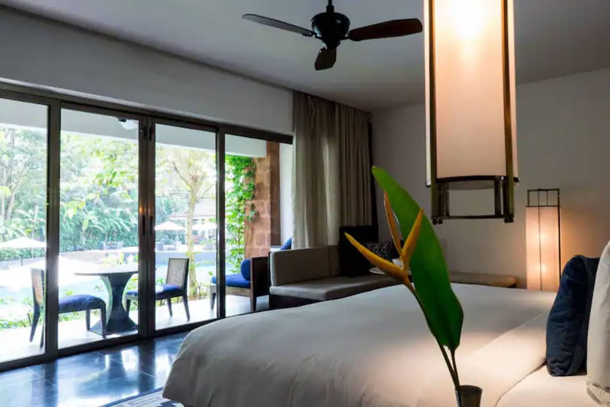A modern hotel room with a large bed, a ceiling fan, a view of the outdoors, and a dining area visible through the glass doors.