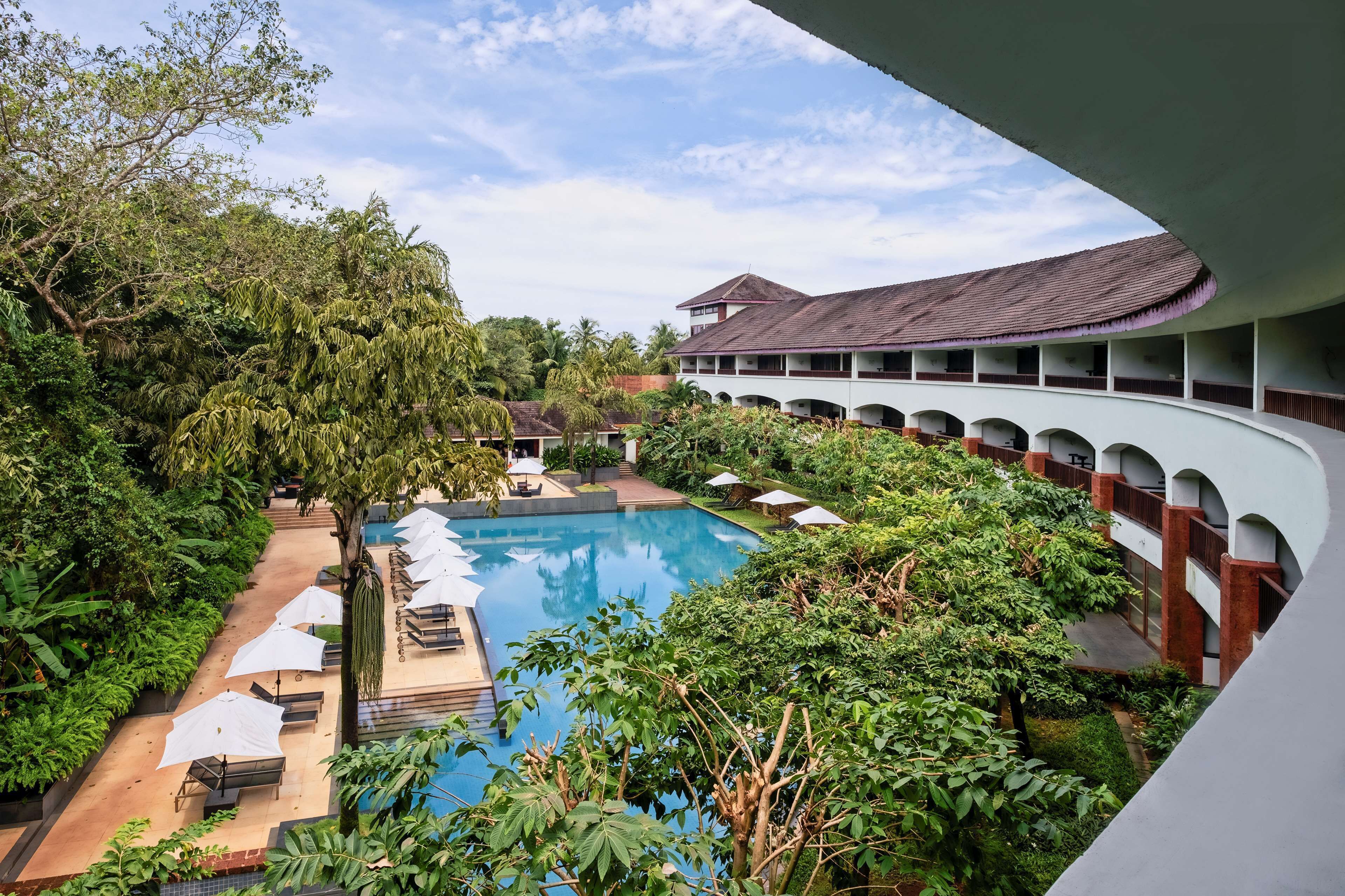 Piscina cercada por vegetação exuberante e um edifício com telhado curvo e várias varandas.