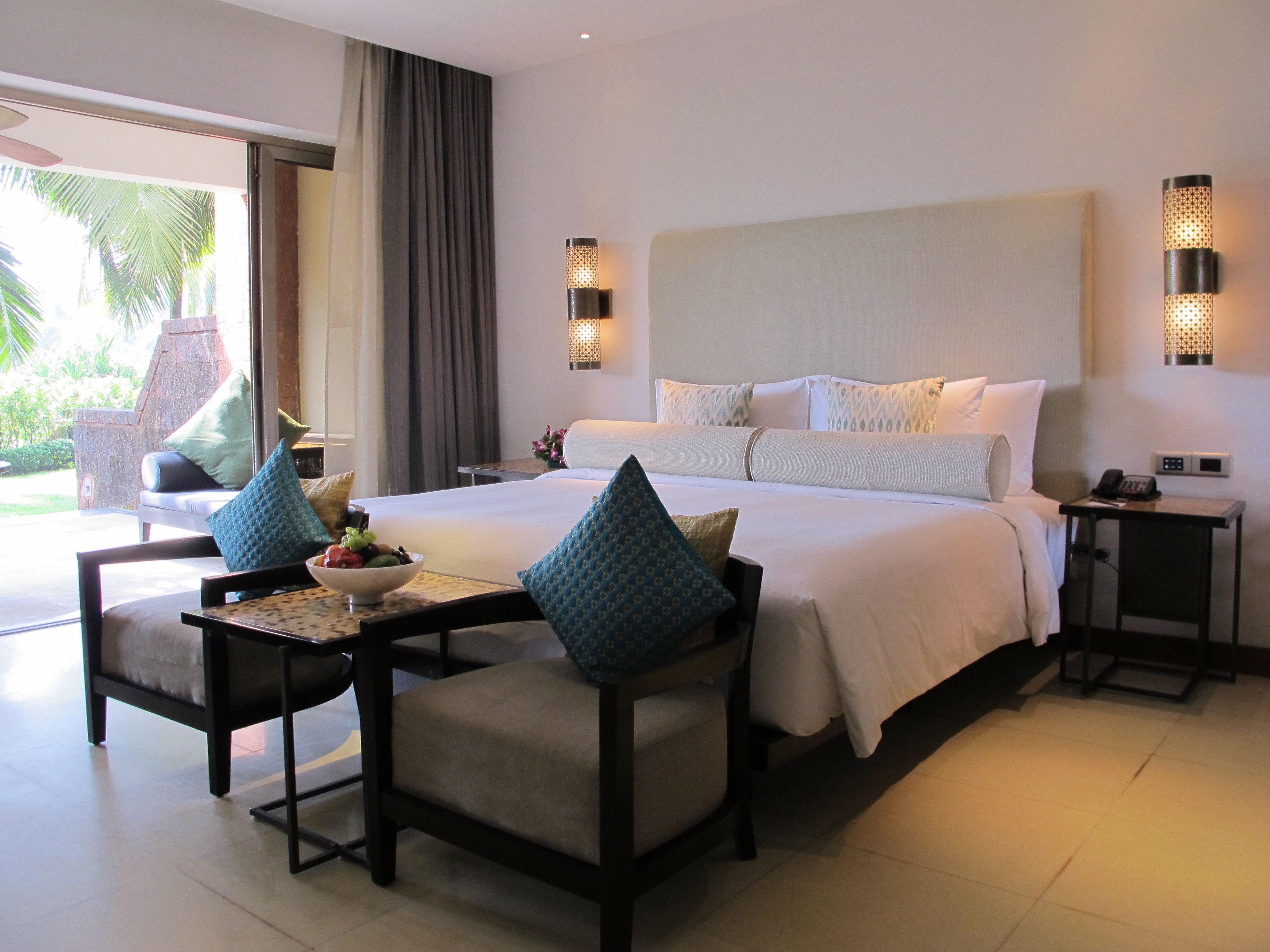 Une chambre d’hôtel avec un grand lit, deux tables de chevet, une petite table sur laquelle se trouve un saladier rempli de fruits et une chaise.