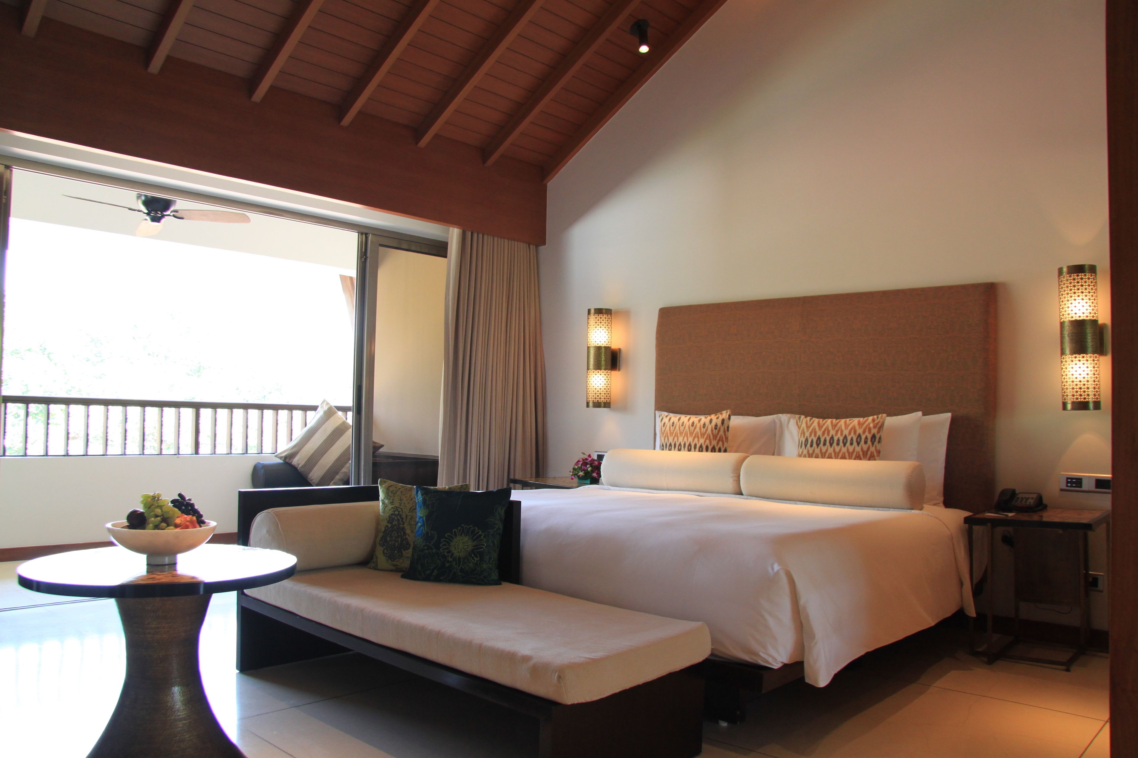 A hotel room with a large bed, a sofa, a small table with a bowl of fruit, and a balcony with a view.