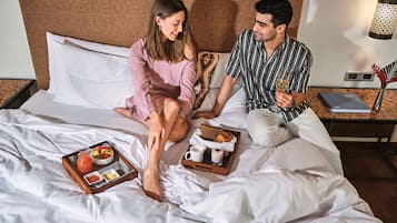 Uma cama com roupa de cama branca, um carrinho com itens de café da manhã e uma mesa de cabeceira com abajur.