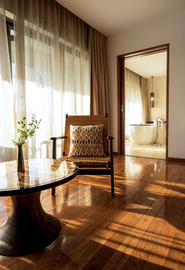 Una habitación con suelo de madera, una mesa redonda con un florero, una silla estampada y vistas a un baño a través de un vano.