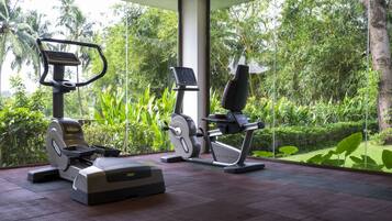 Gimnasio interior con dos bicicletas estáticas y una cinta de correr, rodeado de amplios ventanales con vistas a exuberante vegetación.