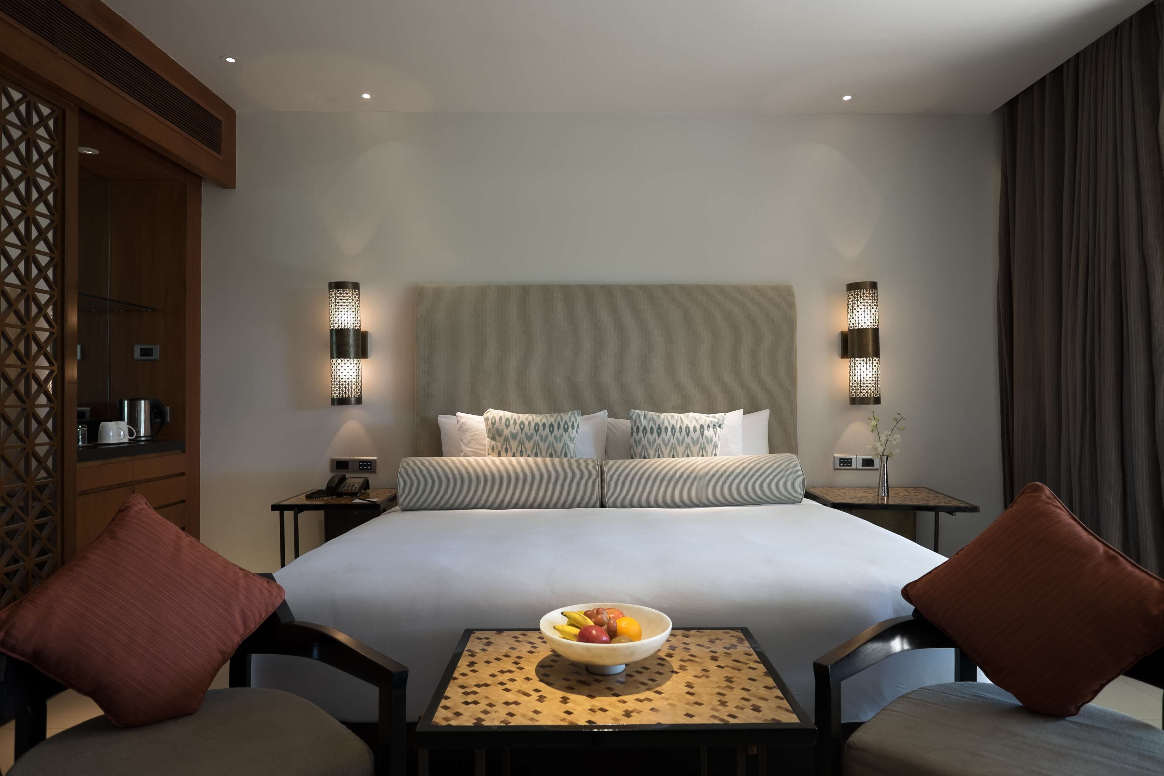 Una habitación de hotel con una cama grande, dos mesitas de noche, una pequeña mesa con un frutero y dos sillas.
