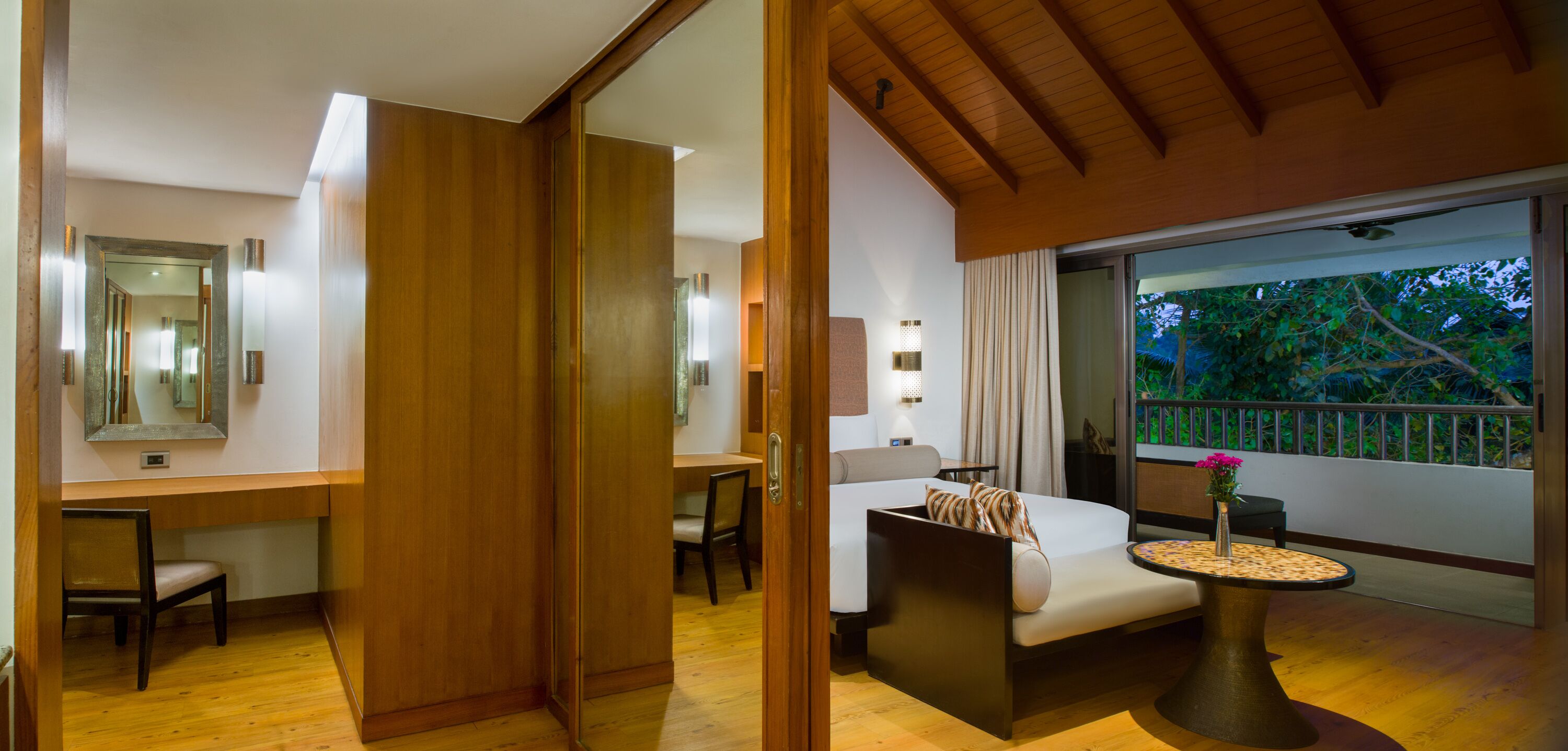 A modern hotel room with a wooden floor, a bed with a pillow, a desk with a chair, a bedside table with a vase, and a window with a view of trees.
