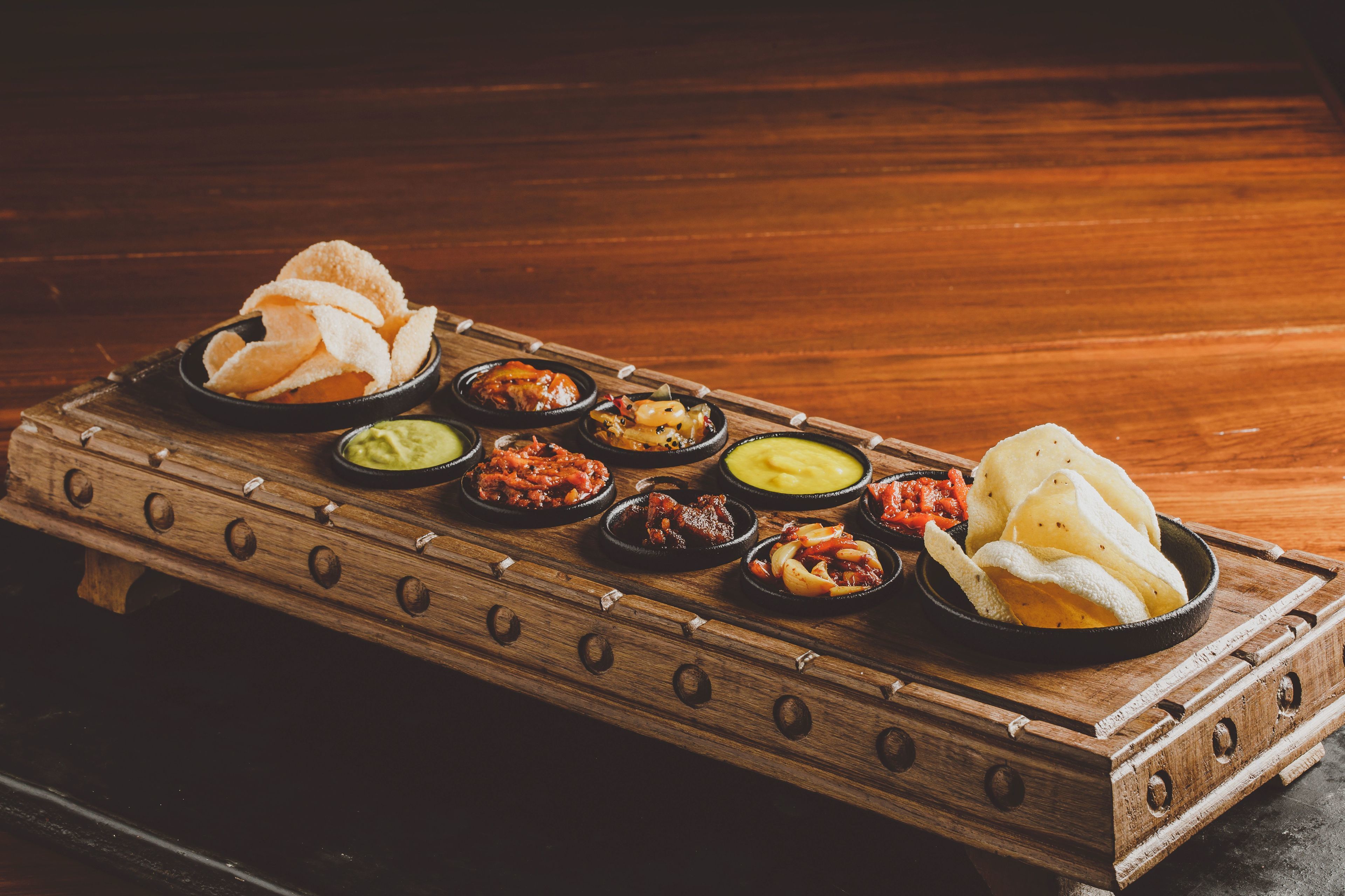 A wooden tray with various dishes including chips, dips, and a bowl of bread.