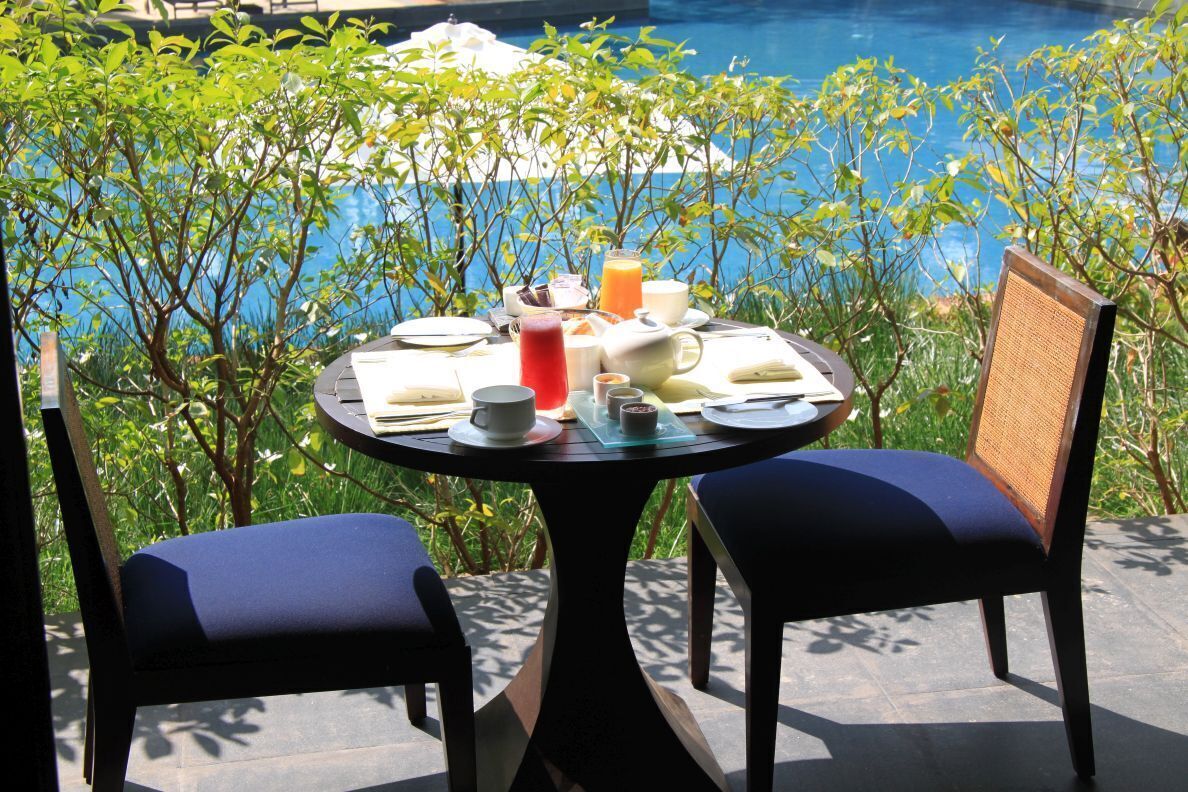 Mesa posta para o pequeno-almoço com vista para uma piscina, rodeada por vegetação exuberante.