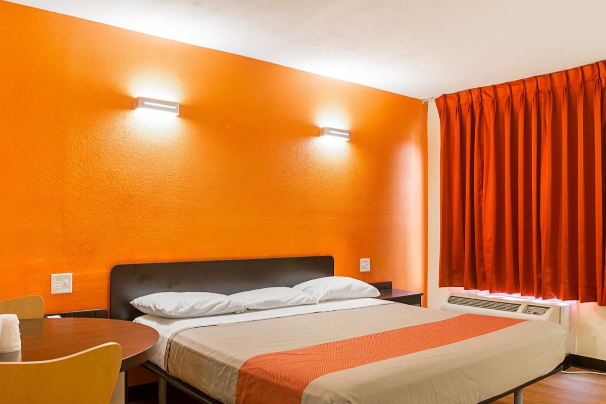 Habitación de hotel con pared naranja, cama con ropa de cama blanca y naranja, un escritorio con una silla y una ventana con cortinas rojas.