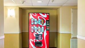 A Coca-Cola vending machine in a hotel corridor with yellow walls and a door on the right.