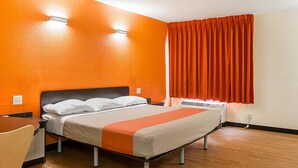 A hotel room with a bed, a wooden floor, an orange wall, and a window with orange curtains.
