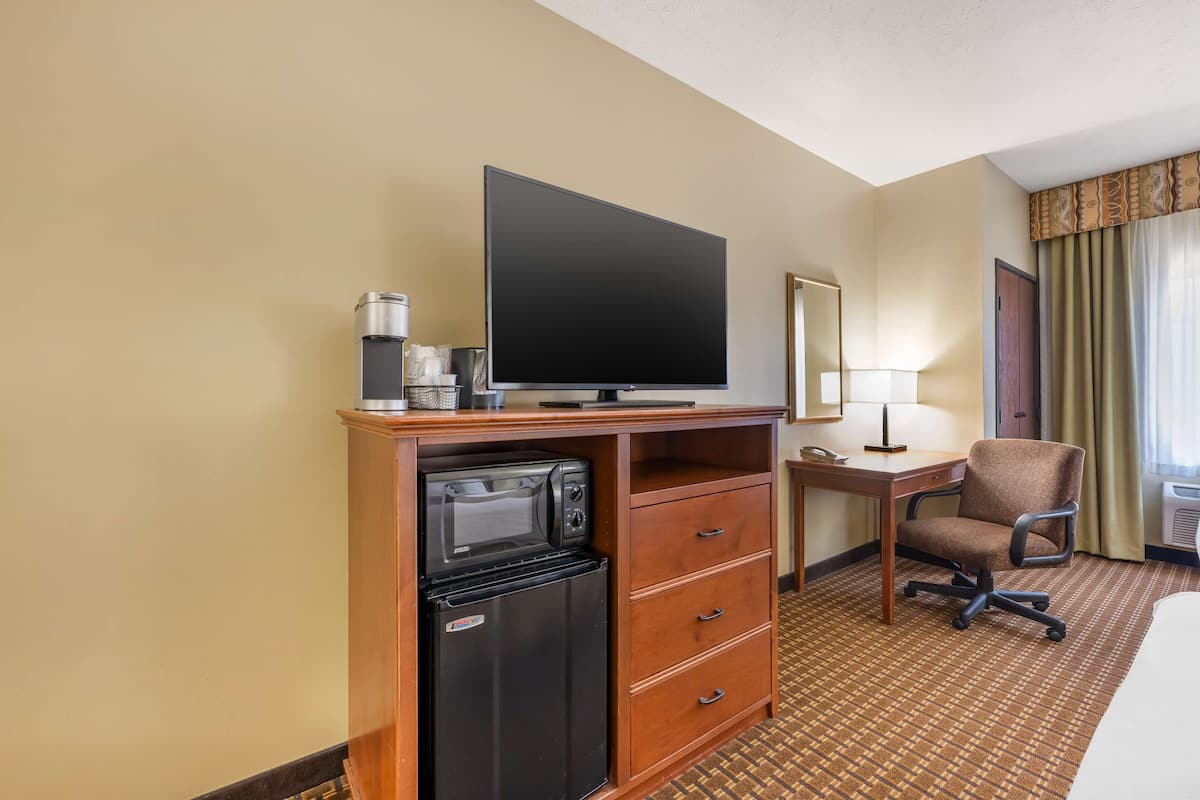 A hotel room with a flat-screen TV, a mini-fridge, a desk with a lamp, a chair, and a bed.