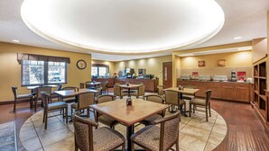 A hotel dining area with multiple tables and chairs, a kitchen in the background, and a large circular light fixture on the ceiling. - Best Western Plus Grand Island Inn & Suites (Grand Island)