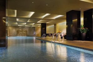 En moderne indendørs swimmingpool med krystalklart vand, en vandfaldsfunktion og et opholdsområde med borde og stole.