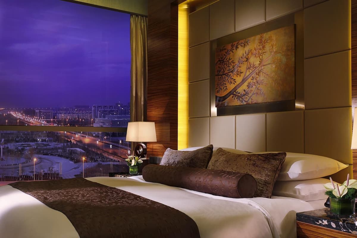 Habitación de hotel con una cama grande, vista nocturna de la ciudad, un cuadro en la pared y una mesita de noche con una lámpara y un florero.