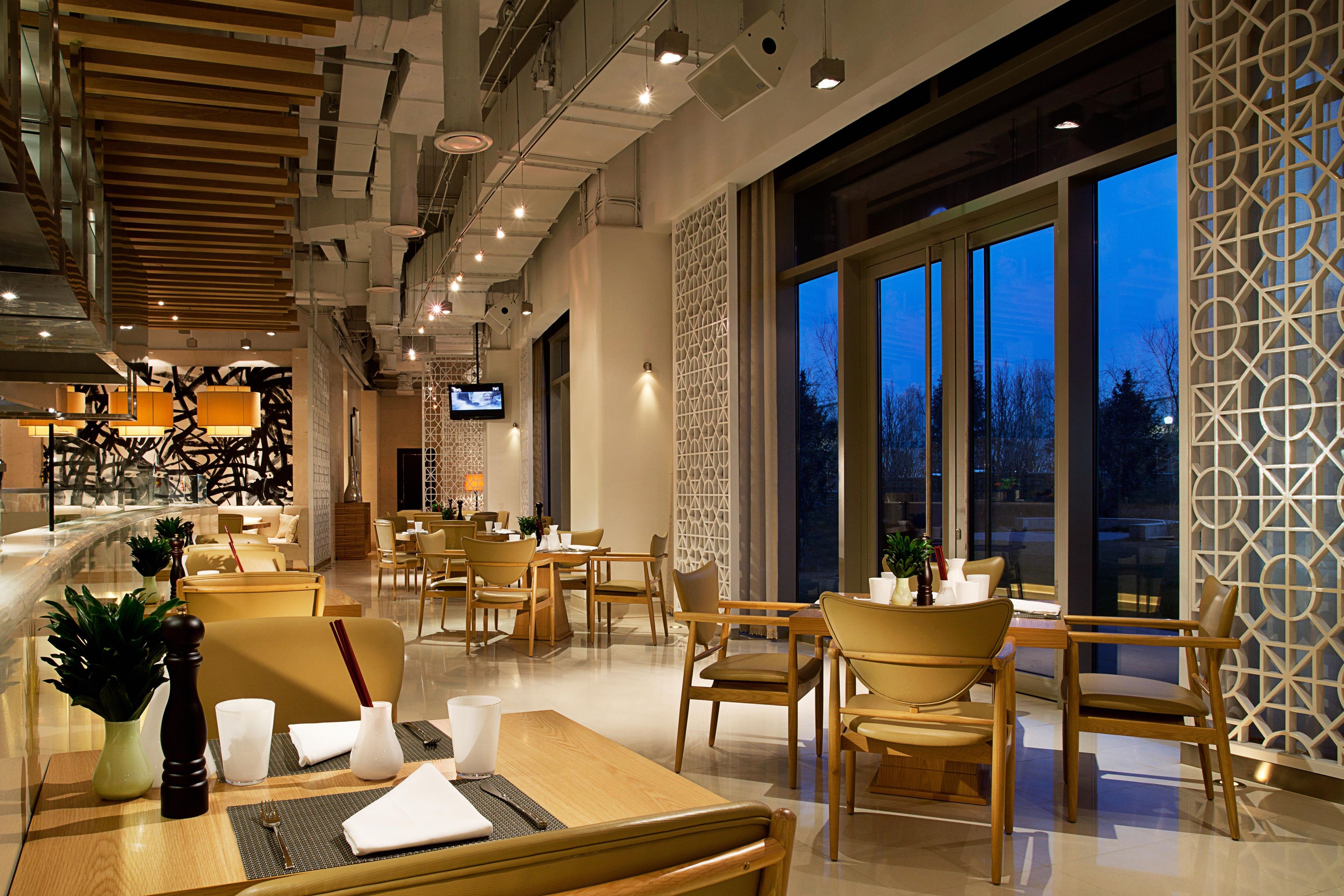 A modern dining area with tables and chairs, a television screen, and large windows.