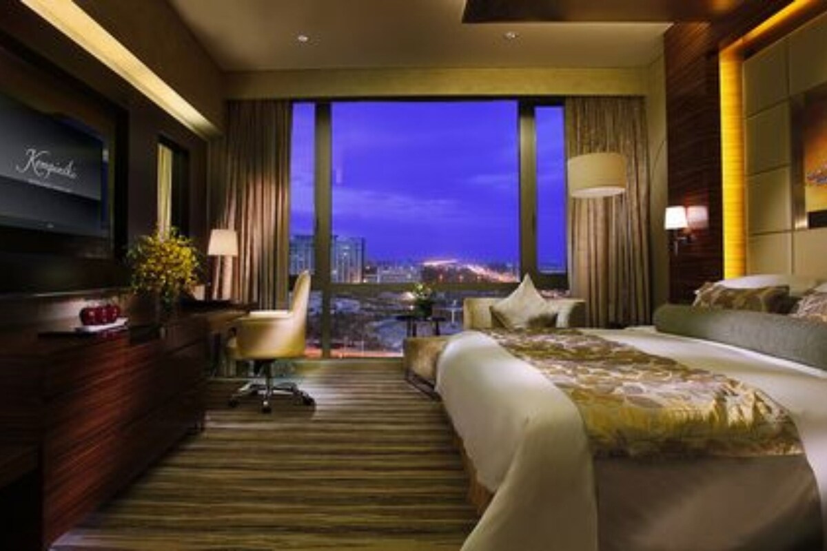 A hotel room with a large bed, a desk with a TV, and a view of the city at night.