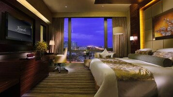 Habitación de hotel con una cama grande, un escritorio con televisor y vistas de la ciudad de noche.