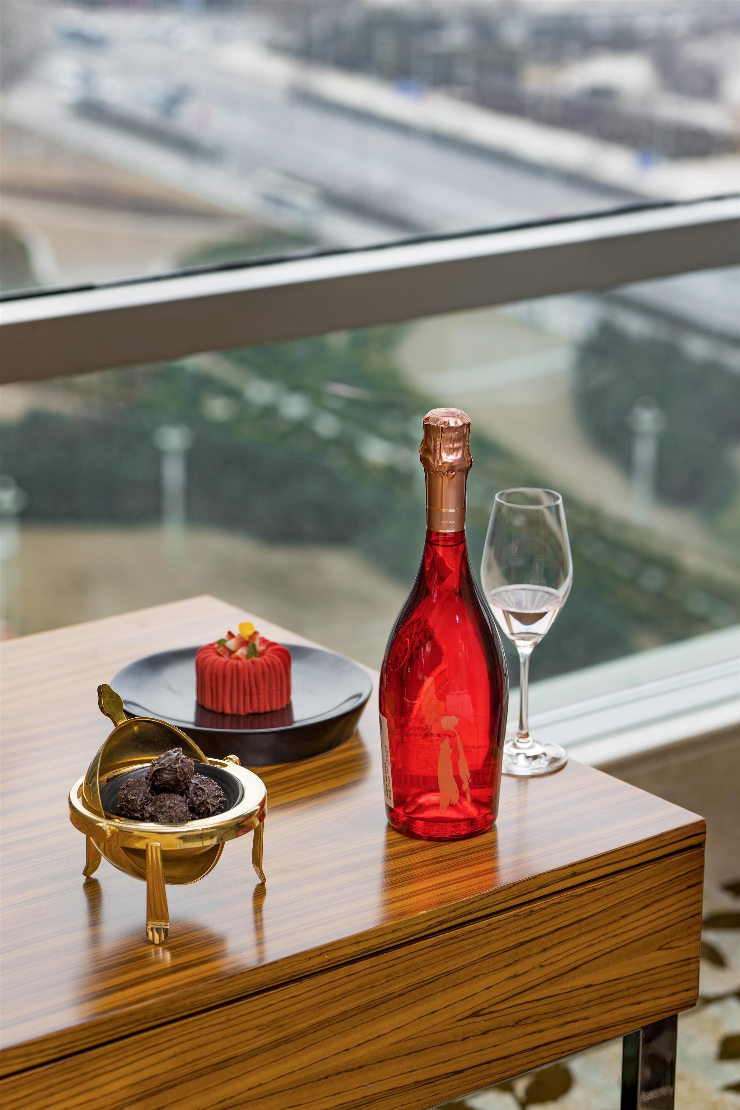 Una mesa con una botella de vino tinto, una copa y un postre sobre una superficie de madera, cerca de una ventana que ofrece vistas a un puente y zonas verdes.