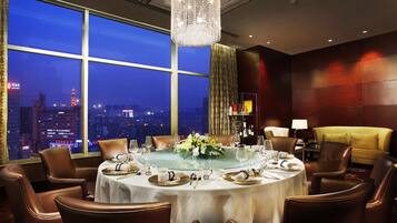 A dining room with a round table set for six, large windows with a city view, a chandelier, and a sofa.