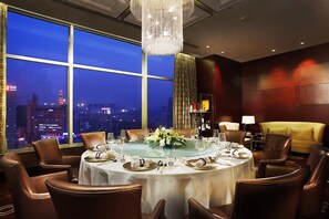 Een eetkamer met een ronde tafel voor zes personen, grote ramen met uitzicht op de stad, een kroonluchter en een bank.
