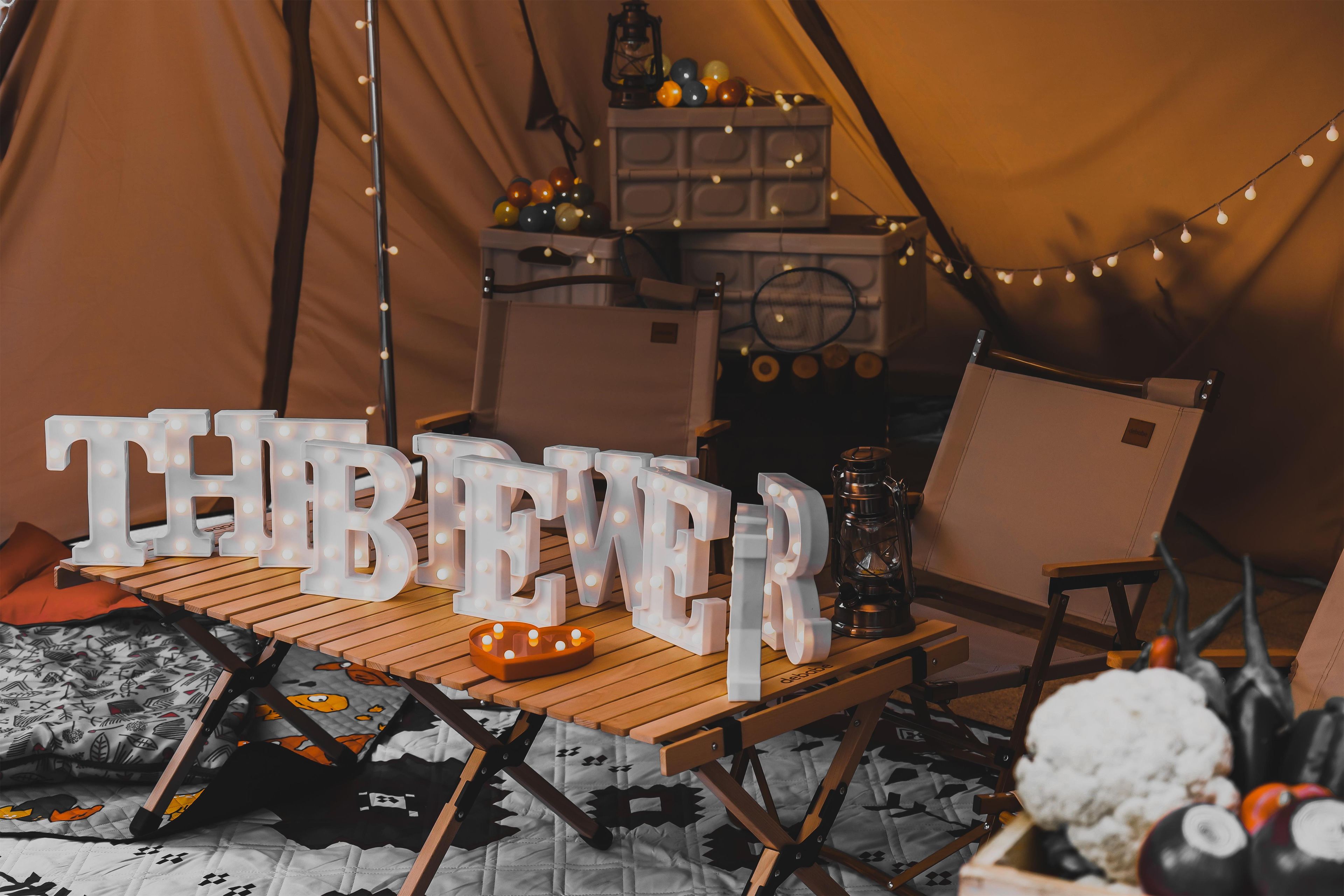 A tent interior with a table displaying illuminated letters spelling 'THERE BEER', a lantern, and a chair.