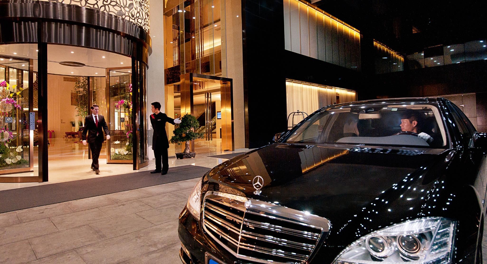 A black luxury car parked in front of a hotel entrance with a concierge and a man in a suit.
