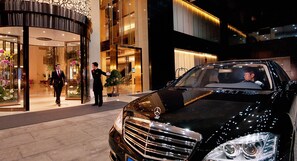 Een zwarte luxeauto geparkeerd voor een hotelingang, met een portier en een man in pak.