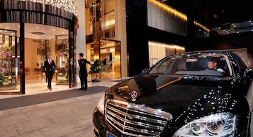 Un coche de lujo negro estacionado frente a la entrada de un hotel, con un botones y un hombre con traje.
