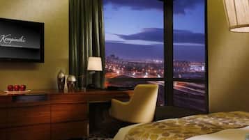 A hotel room with a large window offering a city view at night, a bed, a desk with a lamp, and a television on the wall.