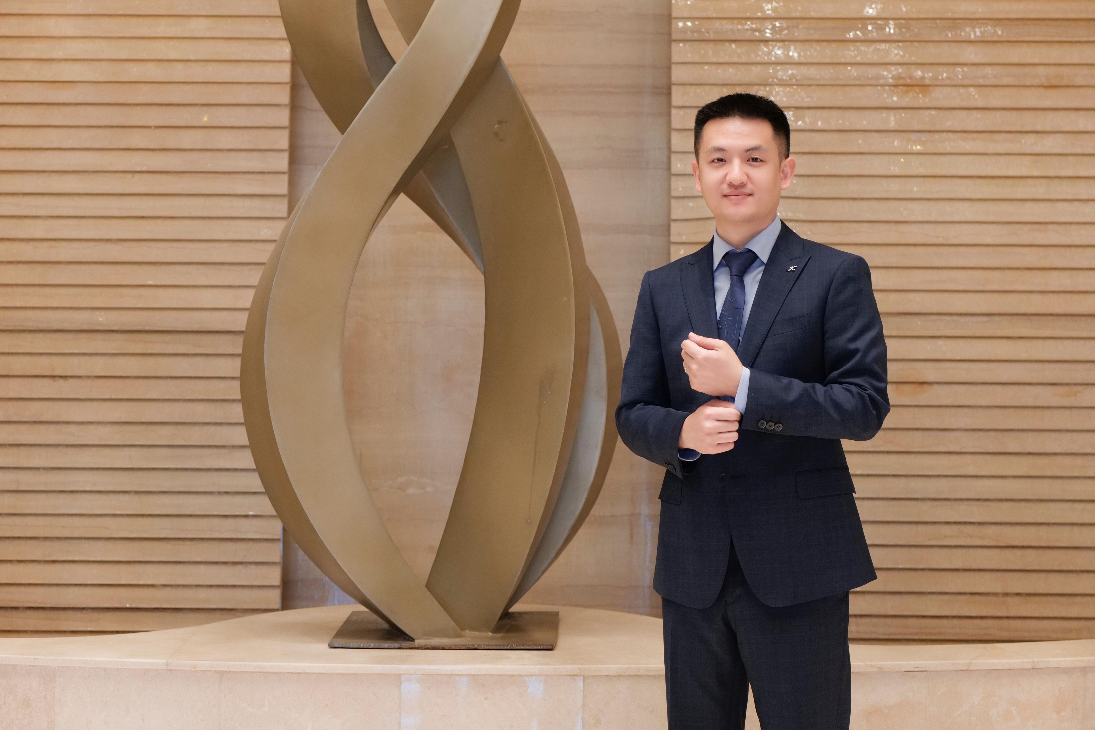 Un hombre con traje de pie junto a una escultura moderna y abstracta en un vestíbulo con paneles de madera.