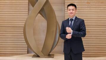 Un hombre con traje de pie junto a una escultura moderna y abstracta en un vestíbulo con paneles de madera.
