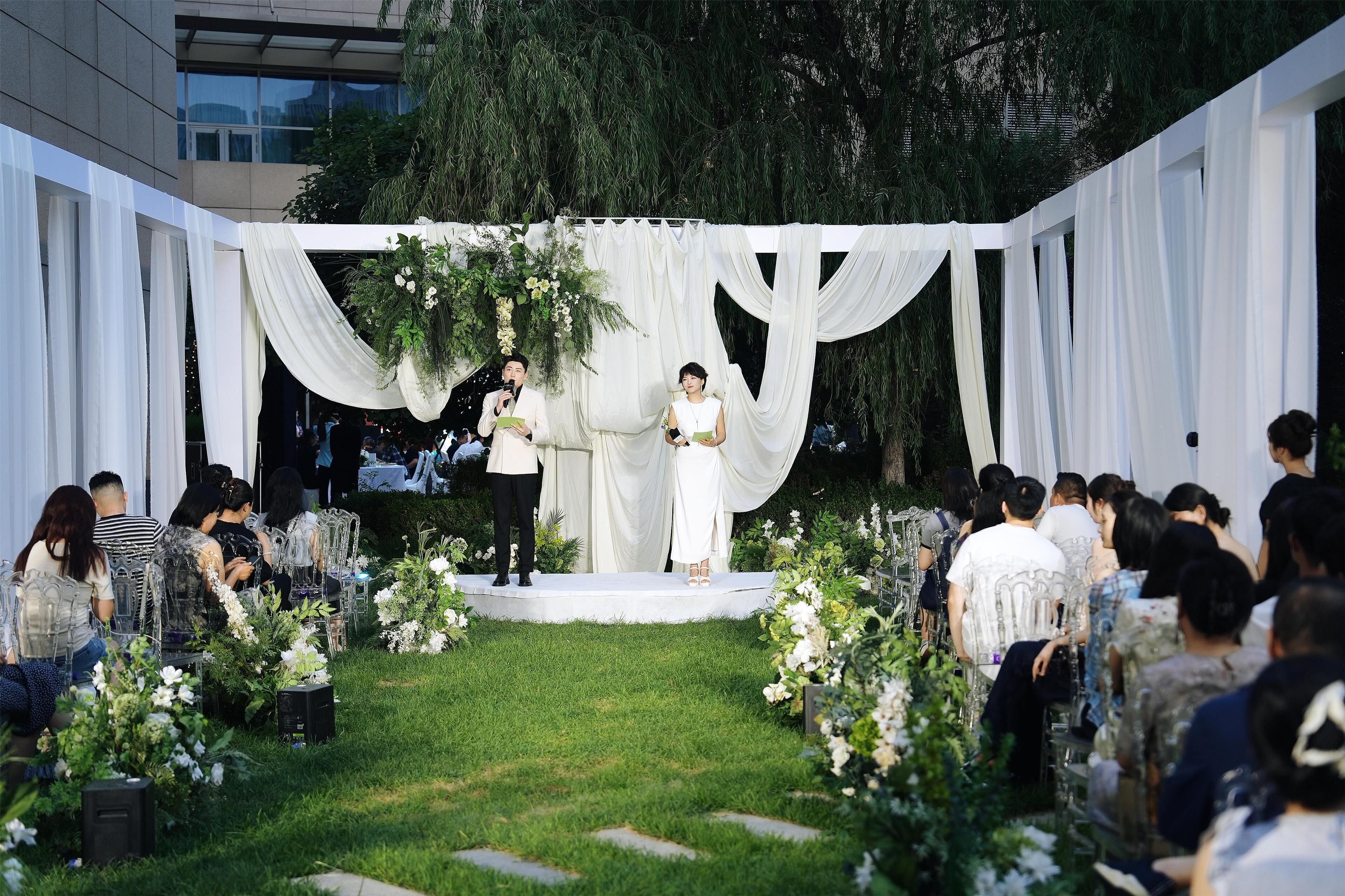 An outdoor wedding ceremony with a stage, guests seated on chairs, and floral decorations.