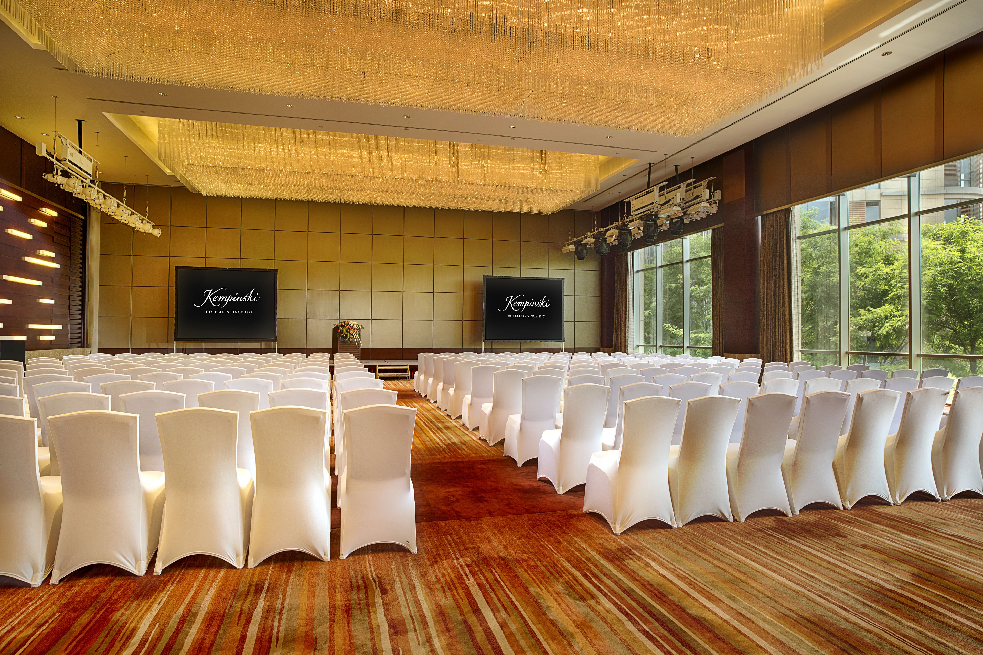 A conference room with rows of white chairs, a large chandelier, and two screens displaying 'Kempinski' branding.