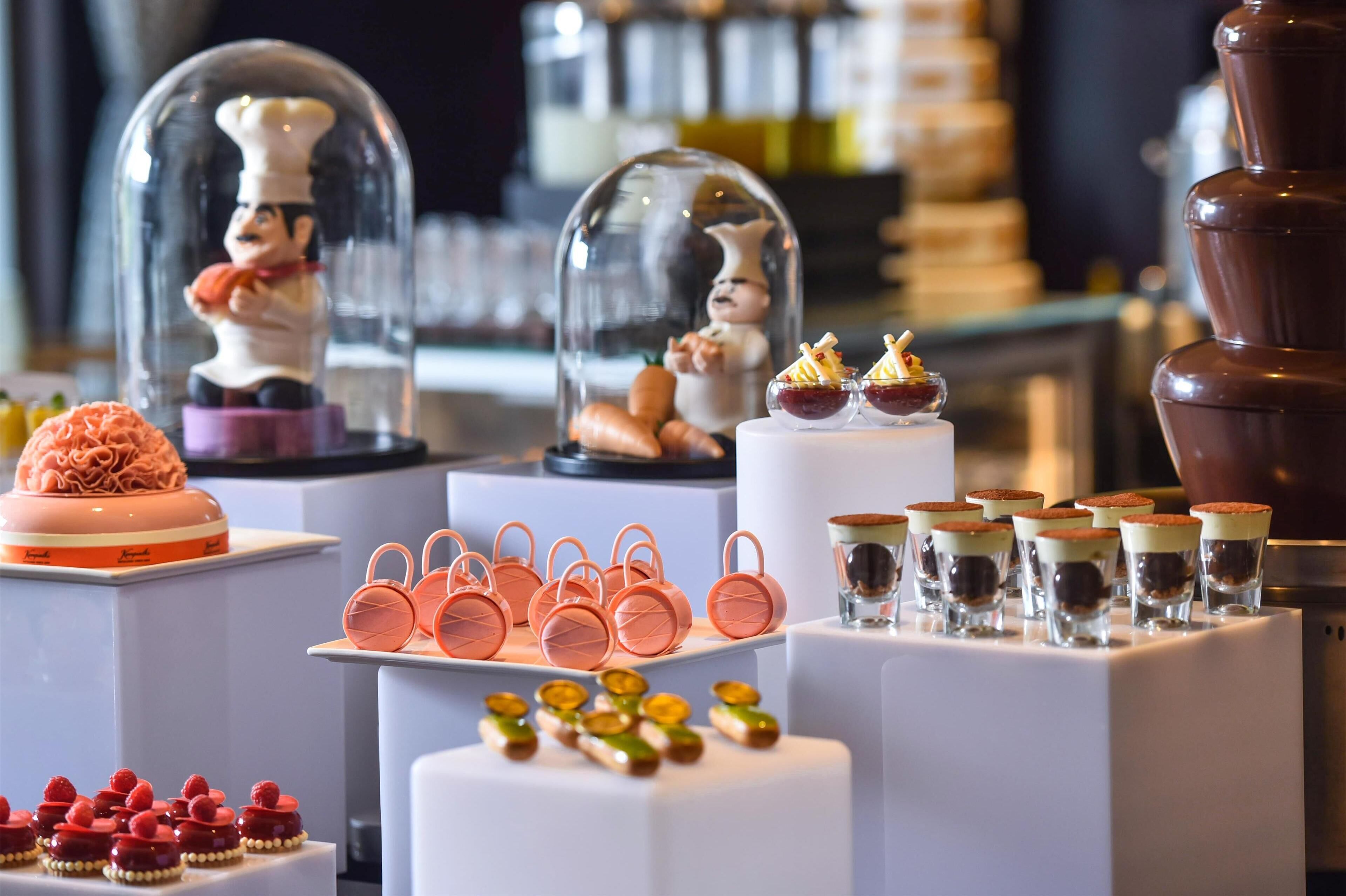 Et dessertdisplay med forskellige kager og søde sager under glasdæksler, blandt andet en lagkage, cupcakes og glas med desserter.